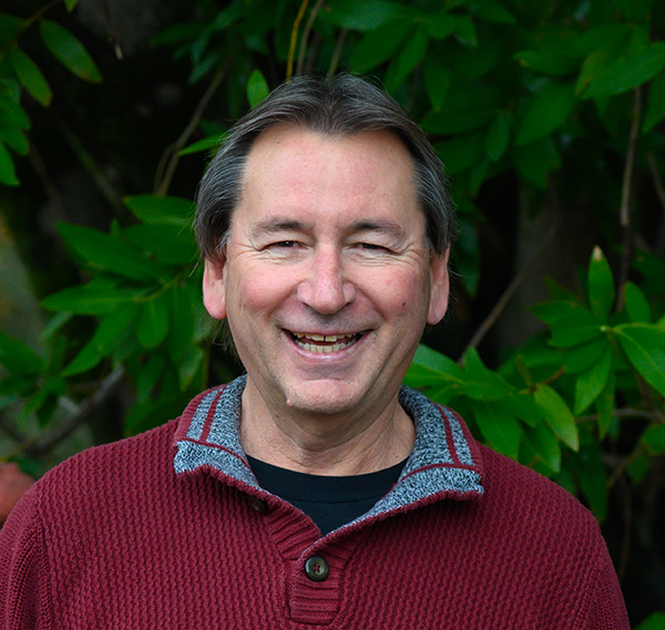 A man in a burgundy sweater smiles in front of a tree.